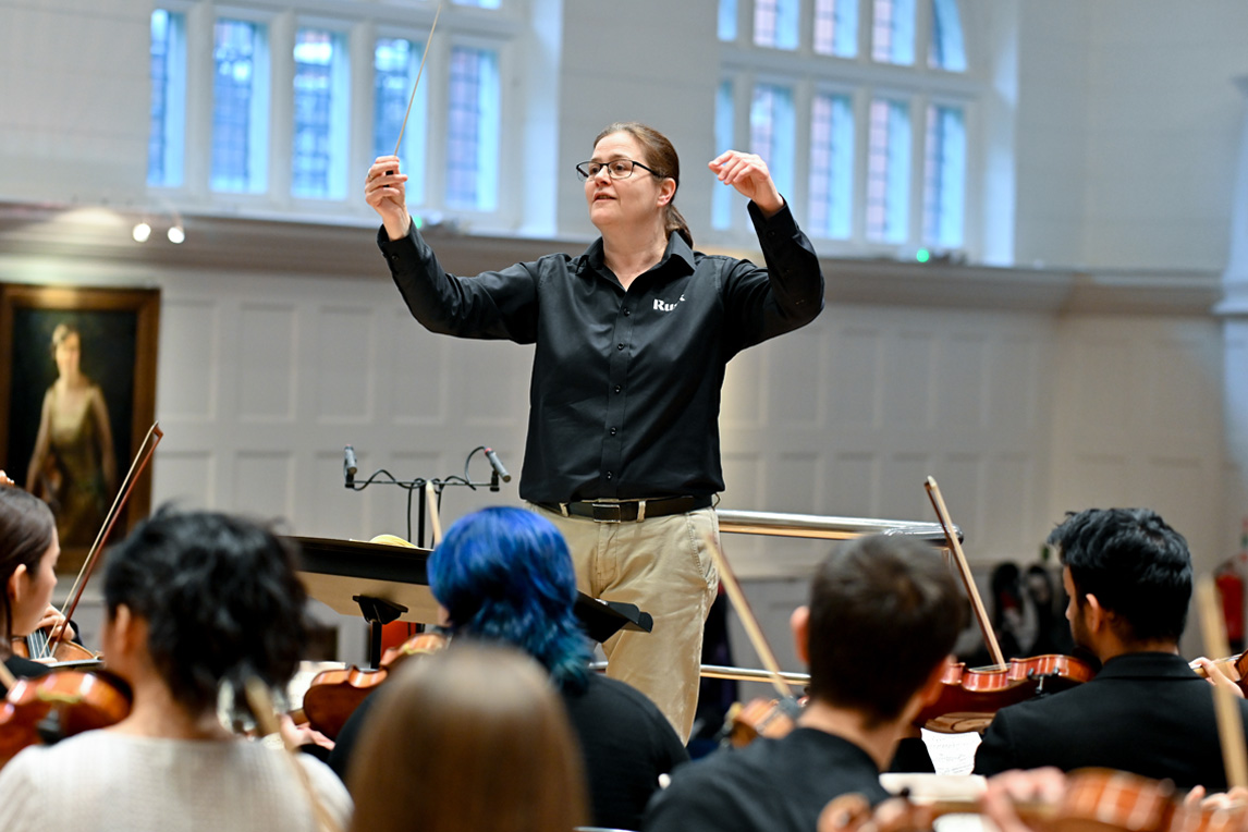 A women wearing a black top and light trousers, leading an orchestral rehearsal.